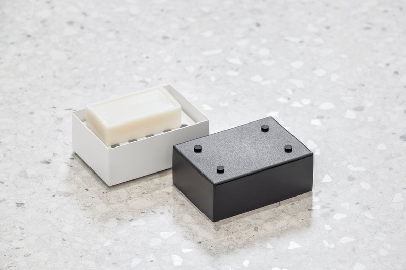 May include: A white soap dish with a bar of soap inside, next to a black soap dish with four rubber feet on the bottom.
