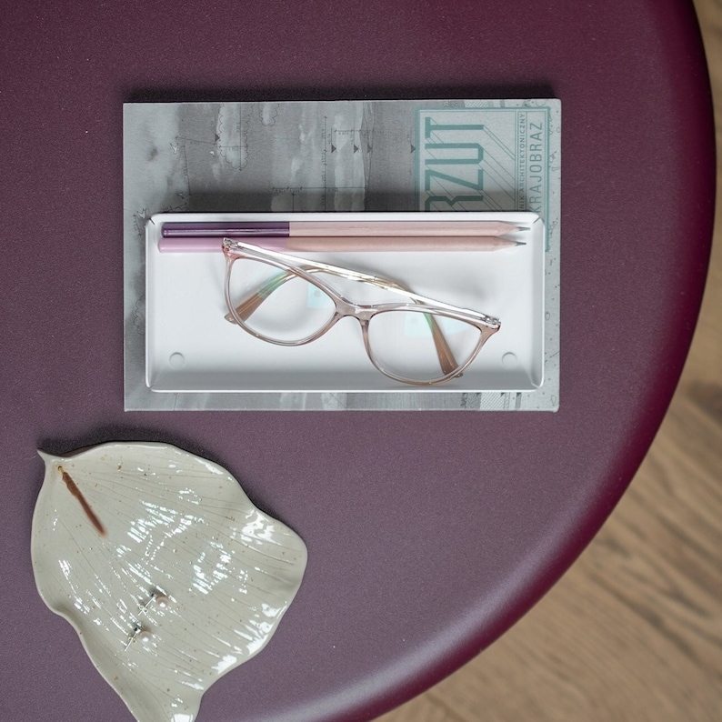 May include: A white rectangular tray with two pencils and a pair of pink-rimmed glasses on a burgundy surface. The tray is sitting on top of a book with the word "RZUT" visible on the cover.