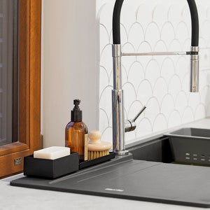 May include: A black metal dish soap dispenser with a clear bottle and a black pump. A wooden brush and a bar of soap are also in the dish rack. The dish rack is sitting on a black countertop next to a black kitchen sink. A chrome faucet with a black rubber hose is in the background.