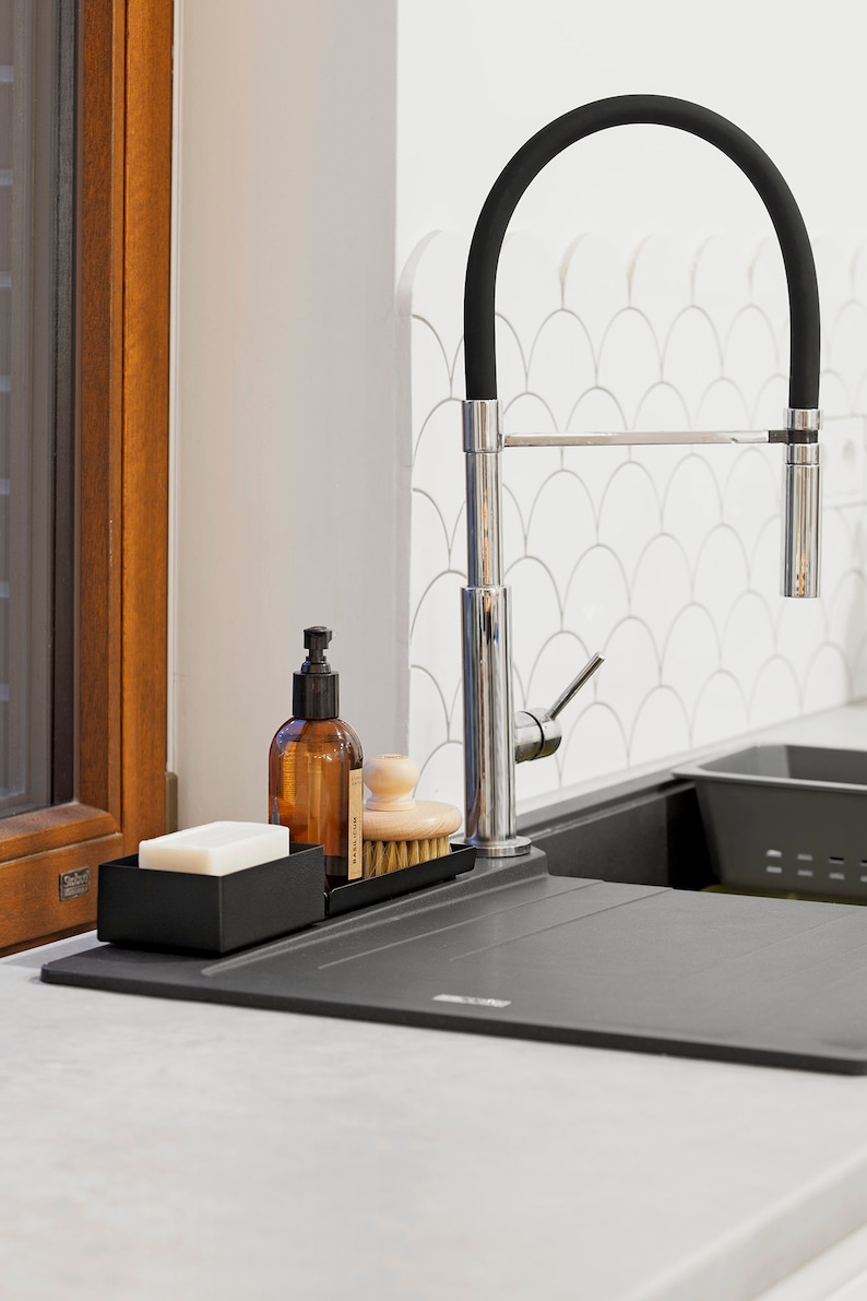 May include: A black metal dish soap dispenser with a clear bottle and a black pump. A wooden brush and a bar of soap are also in the dish rack. The dish rack is sitting on a black countertop next to a black kitchen sink. A chrome faucet with a black rubber hose is in the background.