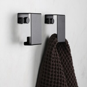 May include: Two matte black metal wall hooks with a flat, rectangular design. Each hook features two circular mounting points on the top section. A dark brown waffle-textured towel hangs from the right hook, showcasing the functional utility.