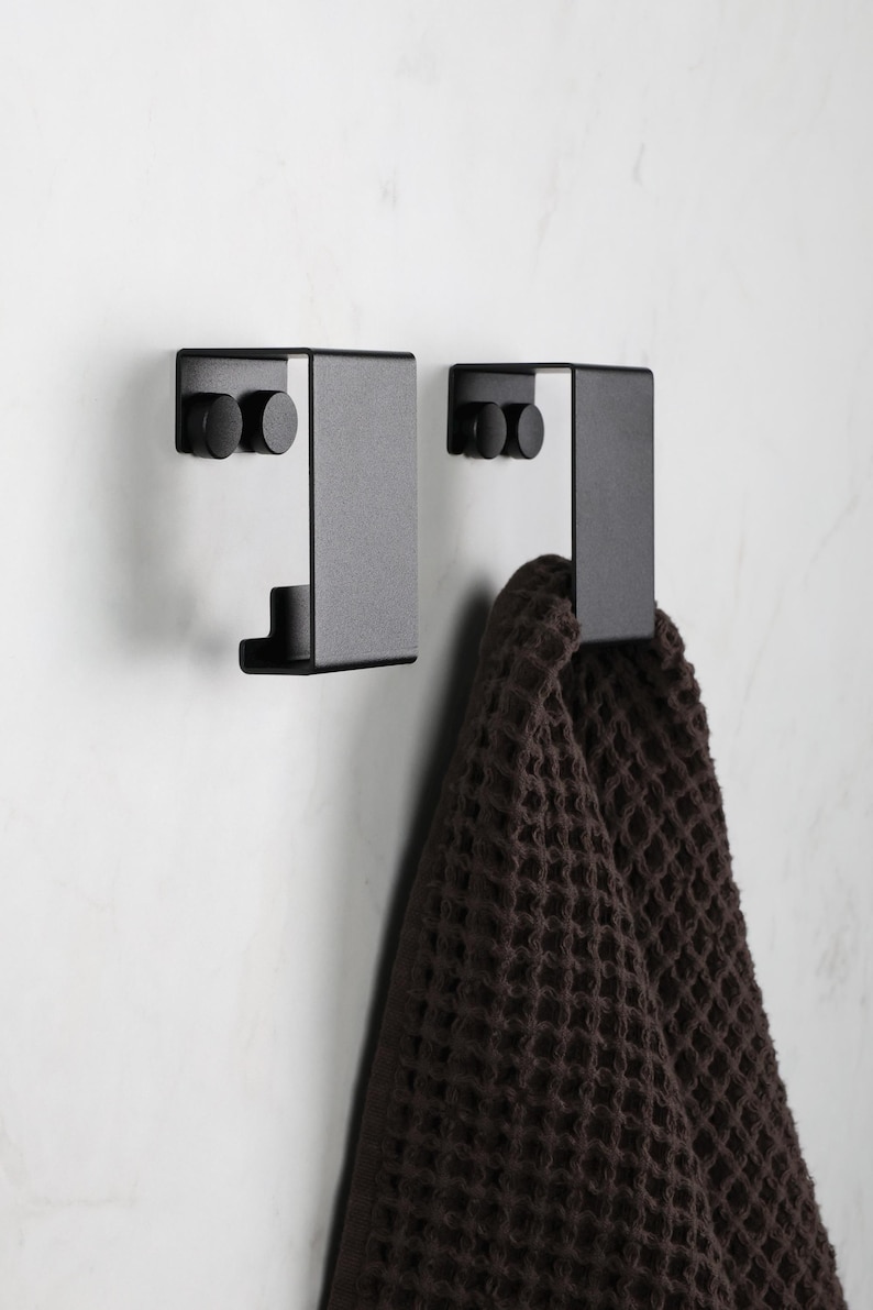 May include: Two matte black metal wall hooks with a flat, rectangular design. Each hook features two circular mounting points on the top section. A dark brown waffle-textured towel hangs from the right hook, showcasing the functional utility.