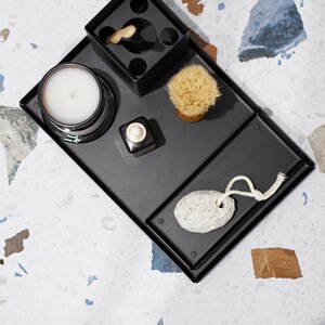 May include: A black metal tray with various items, including a candle, a small bottle, a brush, a pumice stone, and a toothbrush holder. The tray is on a white and gray speckled surface.