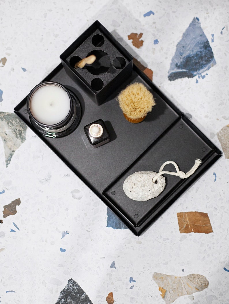 May include: A black metal tray with various items, including a candle, a small bottle, a brush, a pumice stone, and a toothbrush holder. The tray is on a white and gray speckled surface.