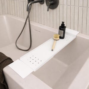 Può includere: Un vassoio da bagno in metallo bianco dal design rettangolare, con una serie di piccoli ritagli circolari. Una spazzola con manico in legno e una bottiglia nera sono posizionate sul vassoio, che si estende sul bordo di una vasca da bagno colorata.