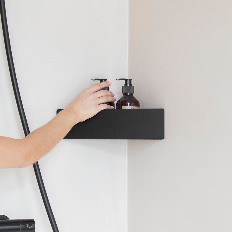 May include: A matte black corner shower shelf mounted on a white wall, featuring a rectangular design with a solid front panel. Two dark amber plastic pump bottles are placed on the shelf surface, providing storage for liquid soap or shampoo products.