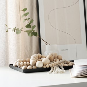 May include: A wooden bead garland with a white tassel hangs on a black tray. The garland is made of natural wood beads in various sizes. The tray is sitting on a white surface.