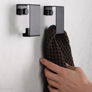 May include: Two matte black metal wall hooks with a rectangular, folded design. Each hook features two circular mounting points on the top section and a curved lower lip for hanging items. A dark brown waffle-textured towel hangs from one hook.