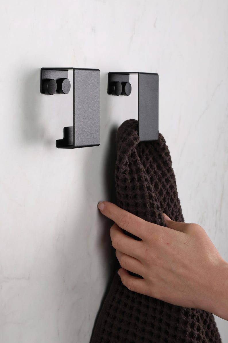 May include: Two matte black metal wall hooks with a rectangular, folded design. Each hook features two circular mounting points on the top section and a curved lower lip for hanging items. A dark brown waffle-textured towel hangs from one hook.