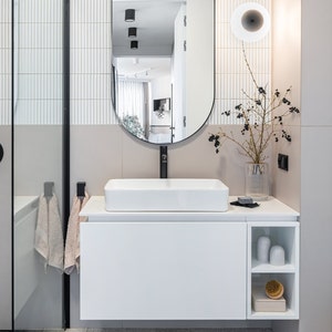 May include: A white rectangular vessel sink sits on a white floating vanity with open shelving. Above, an arched mirror reflects a room. A black faucet, a glass vase with dark berries, a small soap bar, and a wooden brush are on the counter.