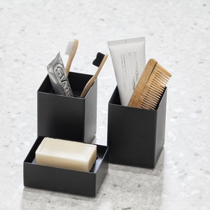 May include: Three black square containers with a white bar of soap in one, a tube of toothpaste and two toothbrushes in another, and a wooden comb in the third.
