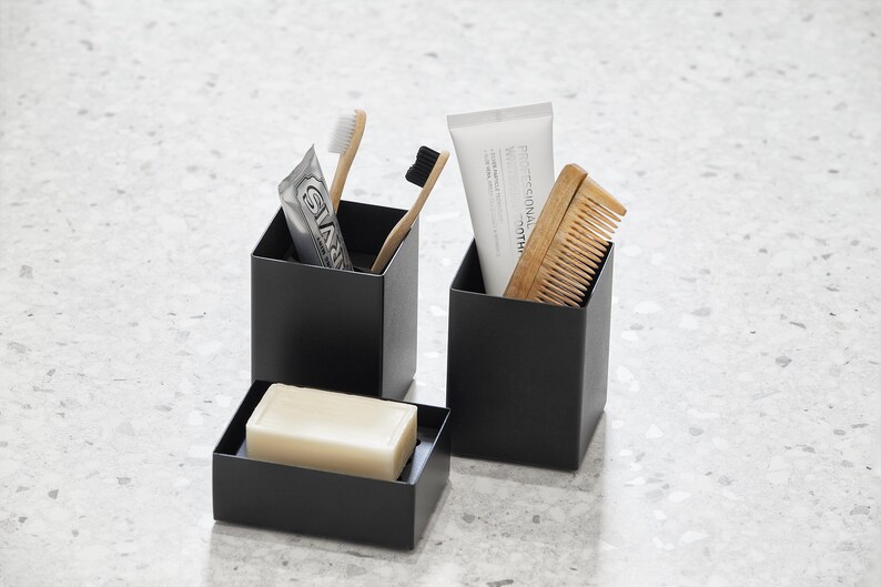 May include: Three black square containers with a white bar of soap in one, a tube of toothpaste and two toothbrushes in another, and a wooden comb in the third.