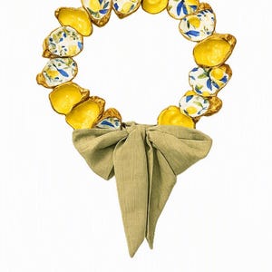 Peut inclure: Une couronne circulaire composée de coquilles d'huîtres individuelles, certaines peintes avec des intérieurs jaune vif et d'autres présentant un motif de citrons bleus et jaunes. Les coquilles sont bordées d'or et finies par un nœud en tissu vert sauge.