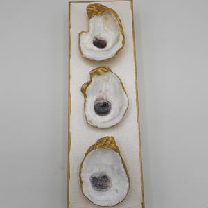 May include: A rectangular canvas art piece featuring three oyster shells. The shells are white with dark centers and gold-painted edges. The canvas has a white background with gold trim, creating a coastal theme.