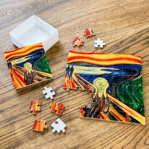 May include: A jigsaw puzzle featuring a reproduction of The Scream painting with swirling orange, yellow, blue, and green colors. The set includes a white box, a square reference card, and several loose puzzle pieces scattered on the surface.