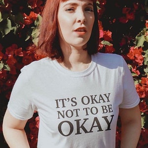 May include: A white t-shirt with black text that reads "It's okay not to be okay". The person wearing the shirt has red hair and is standing in front of a wall of red flowers.