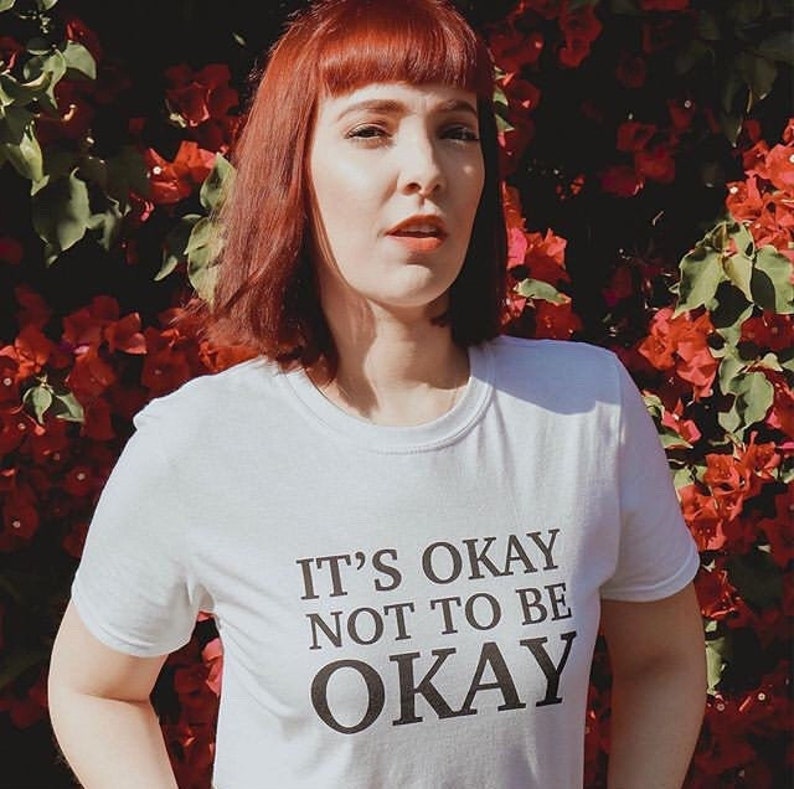 May include: A white t-shirt with black text that reads "It's okay not to be okay". The person wearing the shirt has red hair and is standing in front of a wall of red flowers.