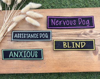 Può includere: Quattro toppe rettangolari in tessuto nero con bordi e testo ricamati. Le toppe riportano le parole Nervous Dog in viola, Assistance Dog in azzurro, Anxious in verde chiaro e Blind in giallo, tutte in un carattere senza grazie.