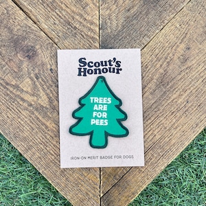 Puede incluir: Una insignia de mérito termoadhesiva para perros, con un diseño de árbol verde y las palabras "TREES ARE FOR PEES" en blanco. La insignia está en una tarjeta beige con el texto "Scout's Honour".