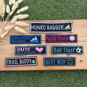 May include: A collection of rectangular embroidered patches with black or blue borders. The patches feature text including MUNRO BAGGER, Adventure Buddy, Wild Child, CUTE BUT PSYCHO, Ball Thief, TRAIL BUDDY, and BEST BOY, alongside icons like mountains, a flower, a heart, a soccer ball, trees, and a smiley face.