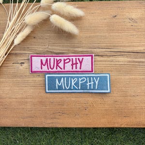 May include: Two rectangular embroidered patches featuring the name MURPHY in capital letters. One patch has a pink background with dark pink stitching, and the other has a blue background with white stitching, both with matching border outlines.