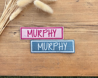 Può includere: Due toppe rettangolari ricamate con il nome MURPHY in lettere maiuscole. Una toppa ha uno sfondo rosa con cuciture rosa scuro, l'altra ha uno sfondo blu con cuciture bianche, entrambe con bordi coordinati.