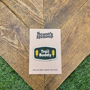 Può includere: Un distintivo al ferro da stiro per cani, con la scritta "Trail Buddy" in un design verde e giallo. Il distintivo è su una carta marrone con le parole "Scout's Honour" in alto. Lo sfondo è un motivo a spina di pesce in legno.