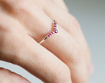 Puede incluir: Un delicado anillo de oro con una fila de pequeñas piedras preciosas rojas en forma de V.