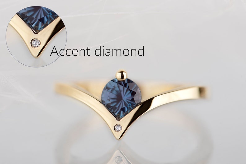 May include: A gold-colored ring with a V-shaped design, featuring a large, round, dark blue gemstone at the center and a small accent diamond on each side. The text "Accent diamond" is visible.