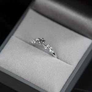 Puede incluir: Un anillo de tono plateado con un grupo de peque&ntilde;as piedras claras de talla redonda dispuestas en un delicado patr&oacute;n floral asim&eacute;trico en la parte superior de la banda, presentado dentro de un joyero gris forrado de terciopelo con tapa.