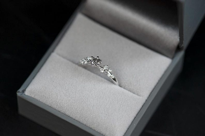 Puede incluir: Un anillo de tono plateado con un grupo de peque&ntilde;as piedras claras de talla redonda dispuestas en un delicado patr&oacute;n floral asim&eacute;trico en la parte superior de la banda, presentado dentro de un joyero gris forrado de terciopelo con tapa.