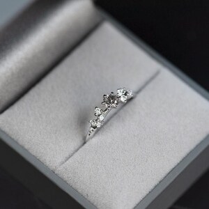 Puede incluir: Un anillo de tono plateado con un grupo de peque&ntilde;as piedras claras de talla redonda dispuestas en un patr&oacute;n asim&eacute;trico y graduado a lo largo de la parte superior de la banda, presentado dentro de un joyero gris forrado de terciopelo.