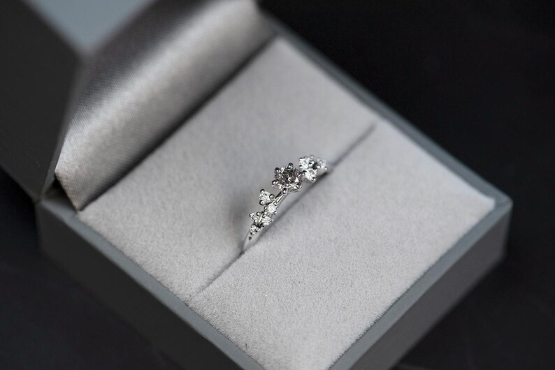 Puede incluir: Un anillo de tono plateado con un grupo de peque&ntilde;as piedras claras de talla redonda dispuestas en un patr&oacute;n asim&eacute;trico y graduado a lo largo de la parte superior de la banda, presentado dentro de un joyero gris forrado de terciopelo.