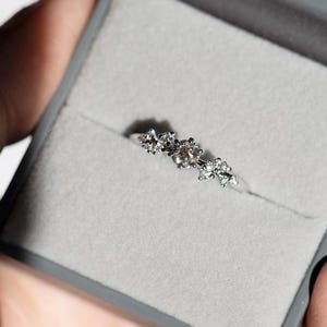 Puede incluir: Un anillo de tono plateado que presenta un grupo de peque&ntilde;as piedras transparentes y facetadas dispuestas en un patr&oacute;n floral en la parte superior de la banda. El anillo se presenta dentro de un joyero gris forrado de terciopelo.