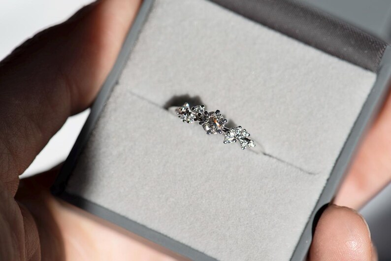 Puede incluir: Un anillo de tono plateado que presenta un grupo de peque&ntilde;as piedras transparentes y facetadas dispuestas en un patr&oacute;n floral en la parte superior de la banda. El anillo se presenta dentro de un joyero gris forrado de terciopelo.