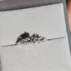 Puede incluir: Un anillo de tono plateado con una piedra preciosa central redonda y transparente, flanqueada por dos grupos m&aacute;s peque&ntilde;os de diminutas piedras transparentes en una banda met&aacute;lica fina, presentado en un joyero gris con interior de terciopelo.
