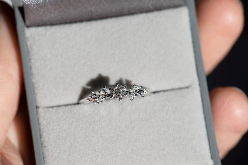 Puede incluir: Un anillo de tono plateado con una piedra preciosa central redonda y transparente, flanqueada por dos grupos m&aacute;s peque&ntilde;os de diminutas piedras transparentes en una banda met&aacute;lica fina, presentado en un joyero gris con interior de terciopelo.