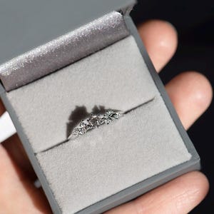 Puede incluir: Un anillo de tono plateado con una piedra central redonda flanqueada por dos piedras m&aacute;s peque&ntilde;as a cada lado, dispuestas en un grupo. El anillo se presenta dentro de un joyero cuadrado de color gris con un forro interior de terciopelo gris claro.