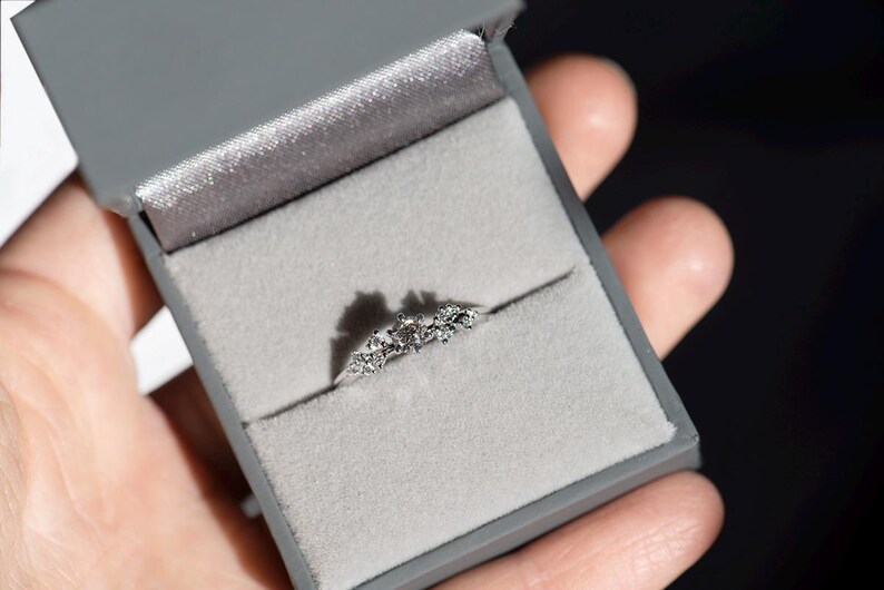 Puede incluir: Un anillo de tono plateado con una piedra central redonda flanqueada por dos piedras m&aacute;s peque&ntilde;as a cada lado, dispuestas en un grupo. El anillo se presenta dentro de un joyero cuadrado de color gris con un forro interior de terciopelo gris claro.
