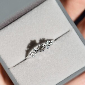 Puede incluir: Un anillo de tono plateado con una piedra central redonda flanqueada por dos grupos de piedras m&aacute;s peque&ntilde;as y claras a cada lado. El anillo se presenta en un joyero cuadrado de color gris con un forro interior de terciopelo gris claro.