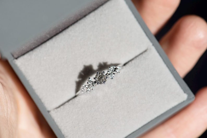 Puede incluir: Un anillo de tono plateado con una piedra central redonda flanqueada por dos grupos de piedras m&aacute;s peque&ntilde;as y claras a cada lado. El anillo se presenta en un joyero cuadrado de color gris con un forro interior de terciopelo gris claro.
