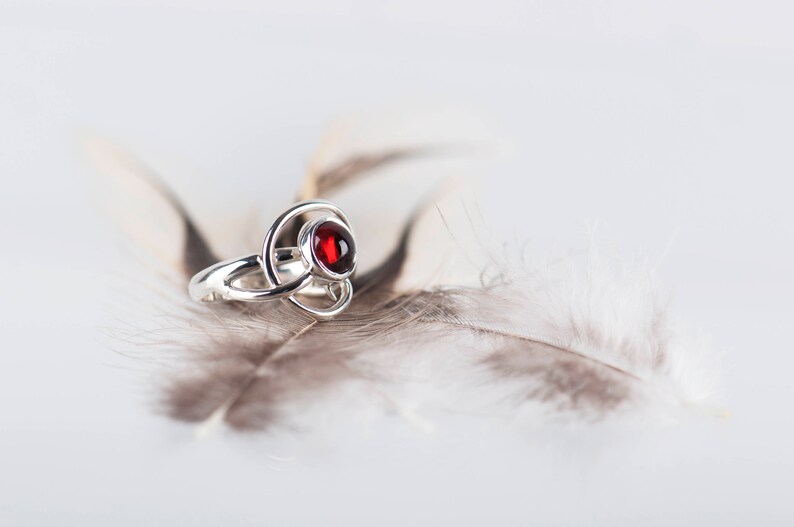 May include: A silver ring with a red gemstone set in a Celtic knot design. The ring is resting on a white feather.