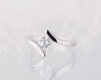 May include: A silver ring with a single, round, clear gemstone set in a simple, modern design. The ring is photographed on a white, feathery background.