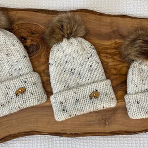 May include: Three white knit beanies with brown faux fur pom poms. Each beanie has a small brown leather patch with a design on it.