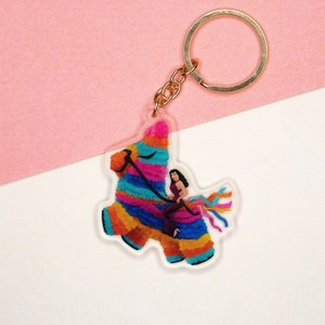May include: A colorful piñata keychain with a gold-colored ring. The piñata is decorated with vibrant blue, orange, yellow, and pink stripes. A woman in a purple dress is riding the piñata, with colorful streamers flowing behind.