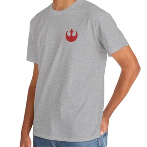 May include: A light gray t-shirt featuring a small red emblem on the chest. The emblem is a stylized symbol. The shirt is made of a soft material and is suitable for casual wear. The person is wearing blue jeans.