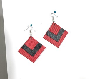 Peut inclure: Une paire de boucles d'oreilles en forme de losange avec une surface rouge texturée et un motif chevron noir. Chaque boucle est suspendue à un crochet en métal argenté avec une petite perle bleue au sommet, formant un design géométrique.
