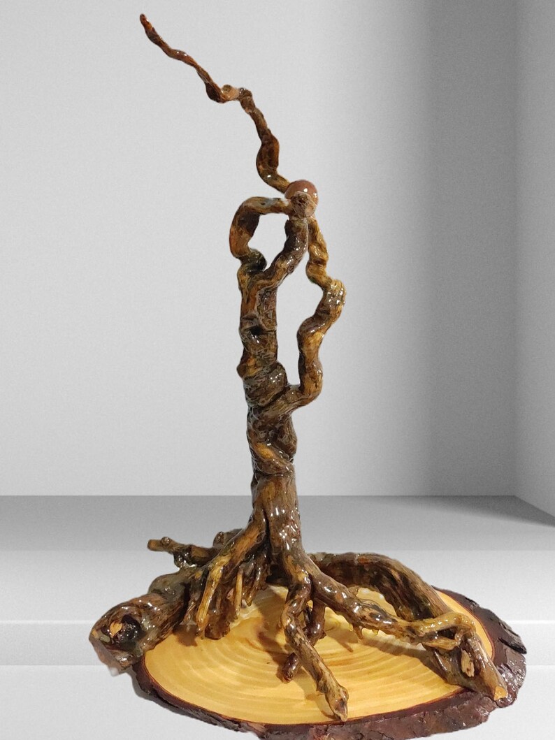 Peut inclure: Une sculpture verticale fabriqu&eacute;e &agrave; partir d'une racine d'arbre brune, polie et torsad&eacute;e, qui s'&eacute;tire vers le haut en une branche fine et courb&eacute;e. La structure racinaire est fix&eacute;e sur une base en bois clair avec des cernes visibles.