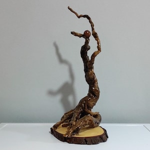 Peut inclure: Une sculpture verticale fabriqu&eacute;e &agrave; partir d'un morceau de bois flott&eacute; torsad&eacute; brun fonc&eacute; poli. Le bois pr&eacute;sente une texture noueuse et organique, fix&eacute;e sur une base circulaire en tranche de bois clair. Une petite sph&egrave;re lisse brun-rouge est en &eacute;quilibre pr&egrave;s du sommet.
