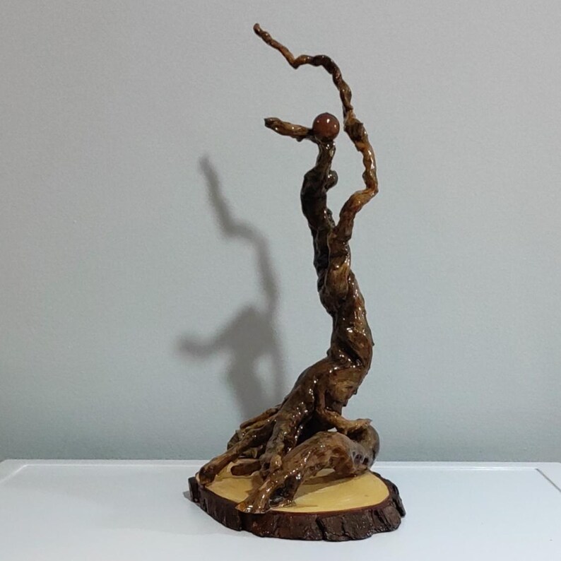 Peut inclure: Une sculpture verticale fabriqu&eacute;e &agrave; partir d'un morceau de bois flott&eacute; torsad&eacute; brun fonc&eacute; poli. Le bois pr&eacute;sente une texture noueuse et organique, fix&eacute;e sur une base circulaire en tranche de bois clair. Une petite sph&egrave;re lisse brun-rouge est en &eacute;quilibre pr&egrave;s du sommet.